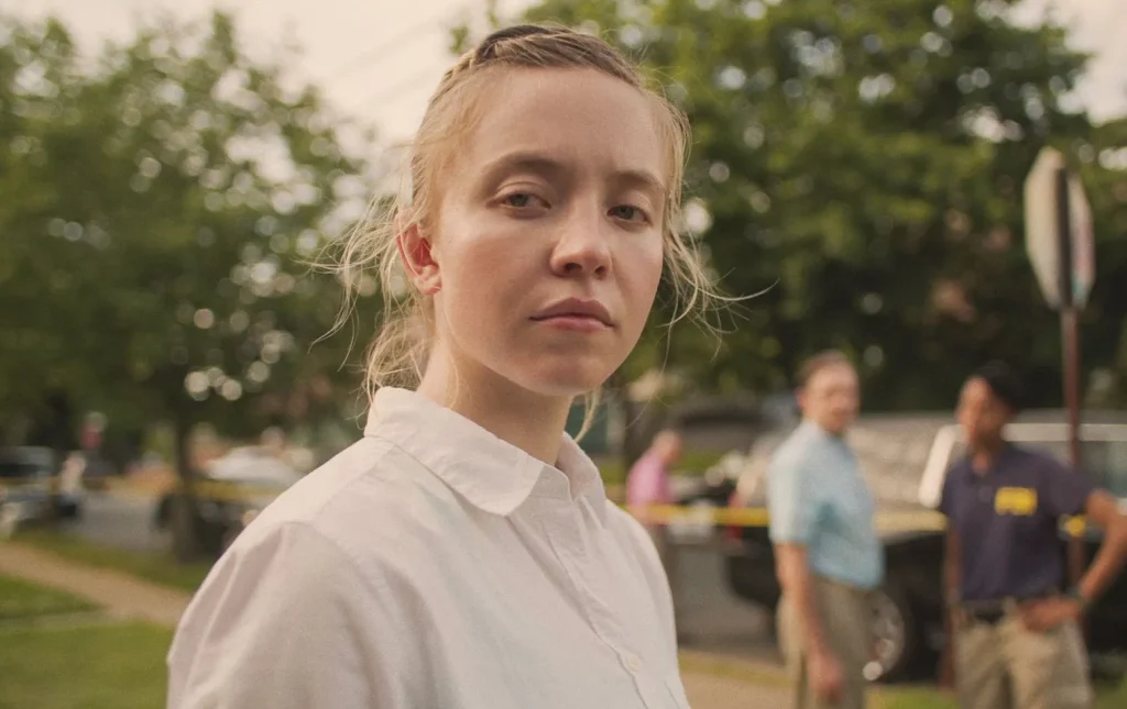 Sydney Sweeney, actriz, interpretando a Reality Winner en la película Reality. Aparece mirando a cámara, y al fondo se ven unos árboles y ados agentes del FBI.