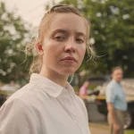 Sydney Sweeney, actriz, interpretando a Reality Winner en la película Reality. Aparece mirando a cámara, y al fondo se ven unos árboles y ados agentes del FBI.