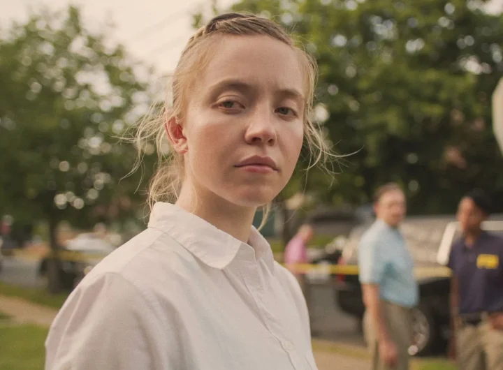 Sydney Sweeney, actriz, interpretando a Reality Winner en la película Reality. Aparece mirando a cámara, y al fondo se ven unos árboles y ados agentes del FBI.
