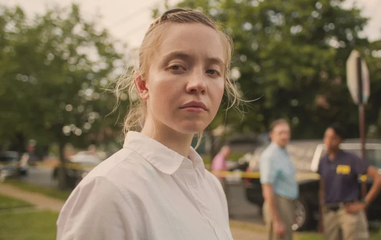 Sydney Sweeney, actriz, interpretando a Reality Winner en la película Reality. Aparece mirando a cámara, y al fondo se ven unos árboles y ados agentes del FBI.